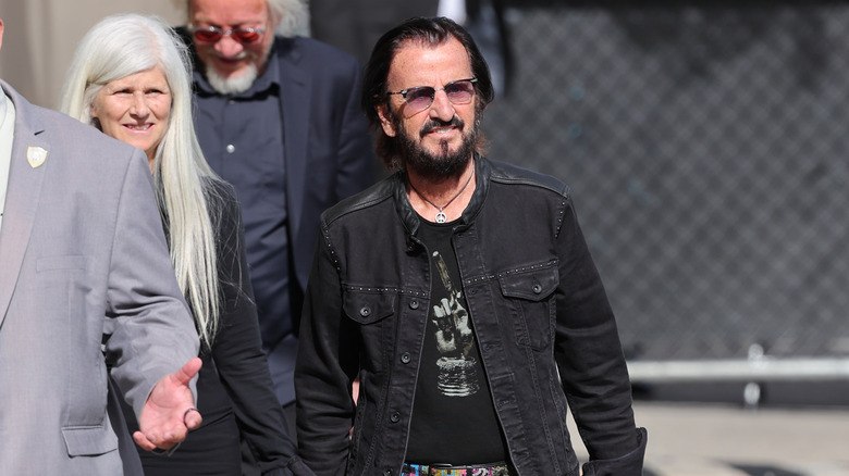 Ringo Starr walking and smiling in an all-black outfit and sunglasses