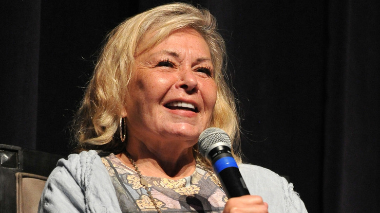 Roseanne Barr holding a microphone onstage in a grey jacket with a patterned shirt underneath