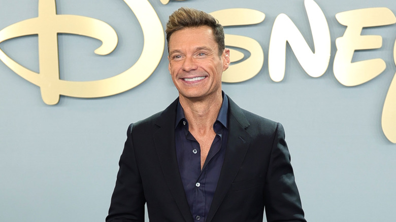 Ryan Seacrest smiling while posing at a Disney event in a black suit