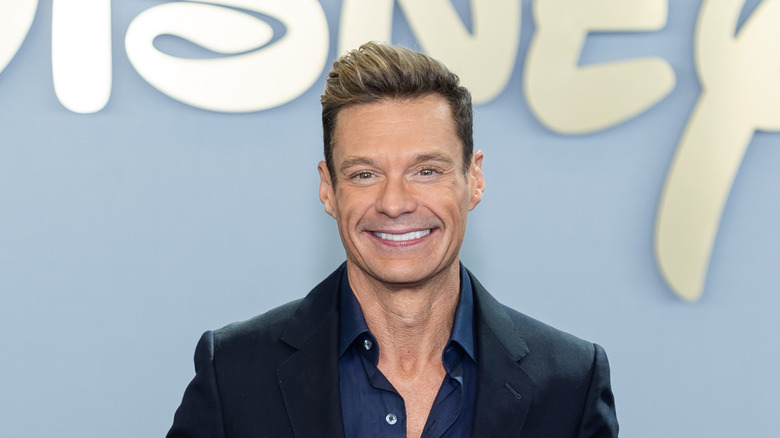Ryan Seacrest smiling while posing at an event in a dark suit