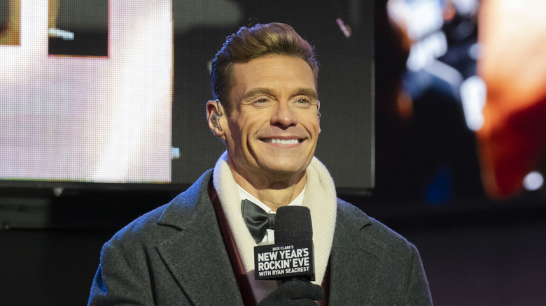 Ryan Seacrest smiling while holding a microphone in a tuxedo and grey overcoat