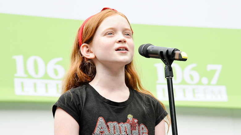 A young Sadie Sink wearing an Annie t-shirt sings in front of a microphone