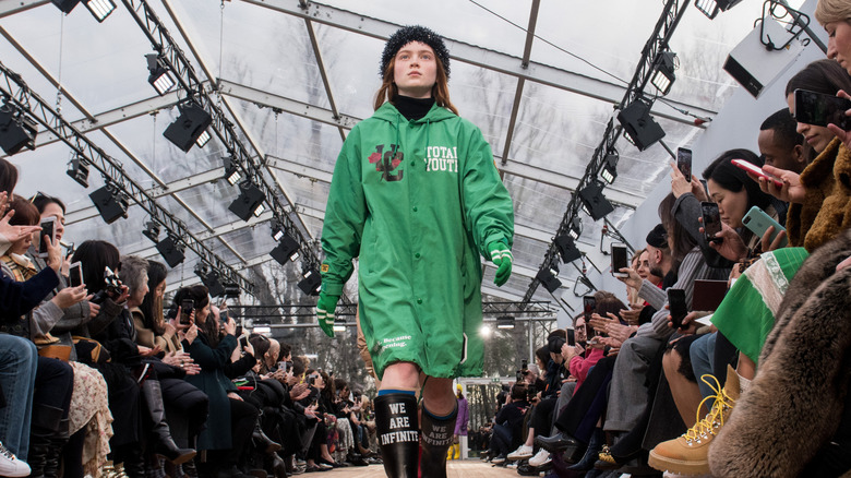 Sadie Sink wearing a green trenchcoat and boots while walking the runway