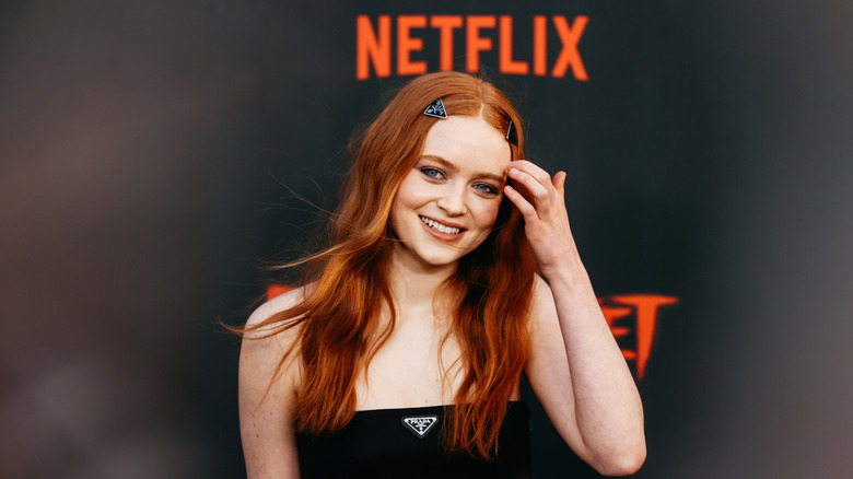 Sadie Sink in a black crop top pushing her hair from her face at a red carpet event