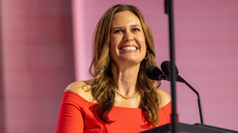 Sarah Huckabee Sanders smiling while standing at a microphone in an off-the-shoulder red dress