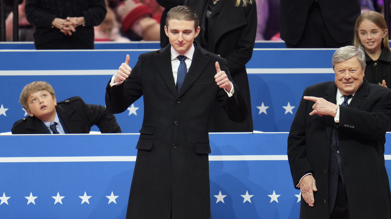 Barron Trump giving two thumbs-up as Viktor Knavs points at him during an indoor inauguration parade at Capital One Arena