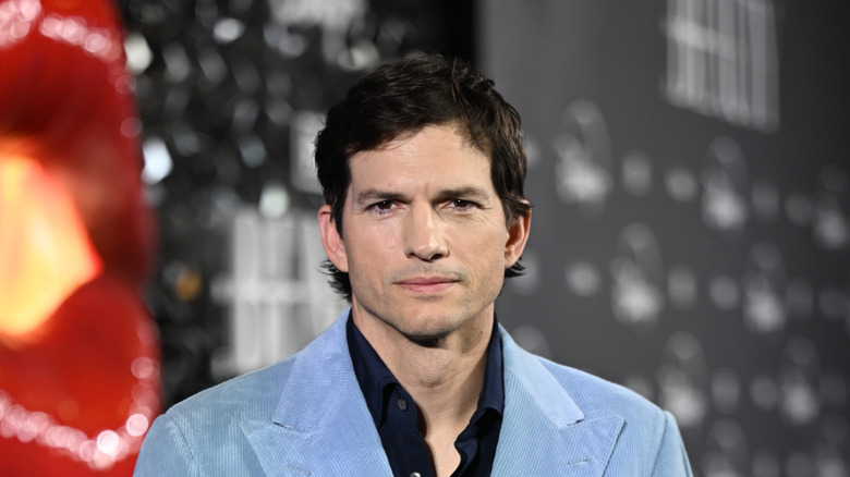 Ashton Kutcher posing for photos in a light blue blazer with a black open-collared shirt underneath