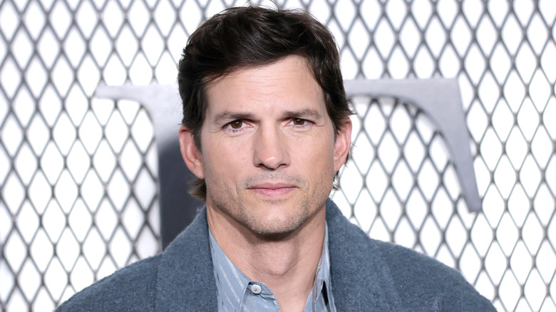 Ashton Kutcher looking at the camera while posing in a grey coat with a light blue striped shirt underneath