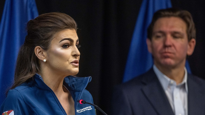 Ron DeSantis looking on as Casey DeSantis talks at a podium on stage