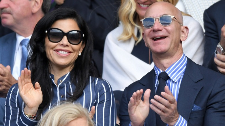 Jeff Bezos and Lauren Sánchez join a crowd and clap their hands while watching a tennis game.
