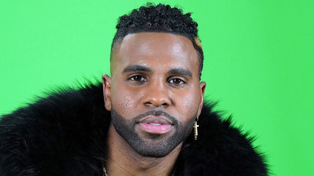 Jason Derulo in a black fur coat and a gold cross earring, in front of a green screen