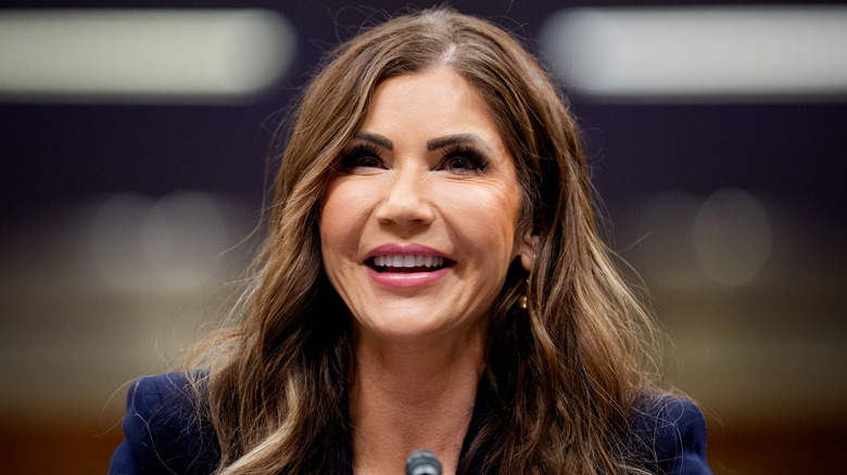Kristi Noem smiling in front of a microphone in a navy blazer