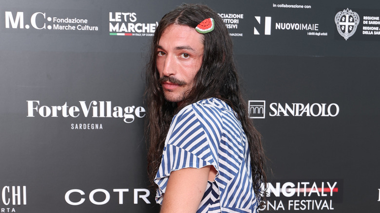 Ezra Miller posing over their shoulder with long hair and a striped dress on the red carpet