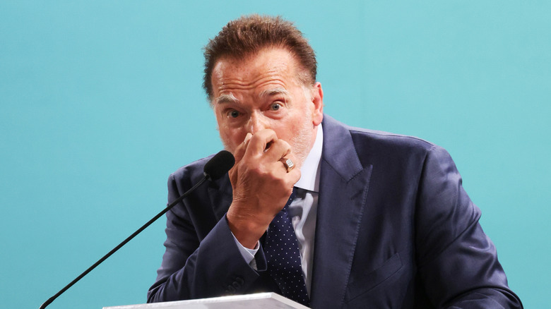 Arnold Schwarzenegger covering his nose and mouth while speaking at a podium while wearing a grey suit, white shirt, and dark blue tie with white polka dots
