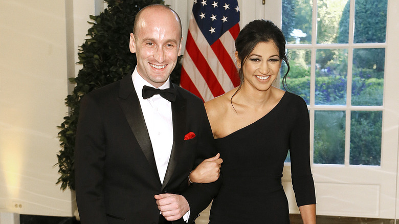 Stephen Miller and Katie Miller walking arm-in-arm and smiling in front of an American flag at the White House