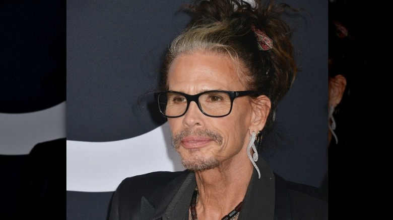 Steven Tyler wearing spectacles on red carpet in a black suit with hair in bun