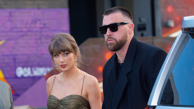 Taylor Swift wearing a thin strap dress walking alongside Travis Kelce in a blue tux and sunglasses