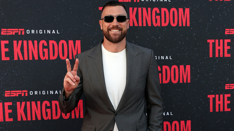 Travis Kelce posing with a peace sign in a grey suit jacket, white shirt, and sunglasses