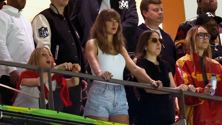 Wearing a white tank top and shimmery shorts, Taylor Swift holds onto a rail while watching a game with other spectators.