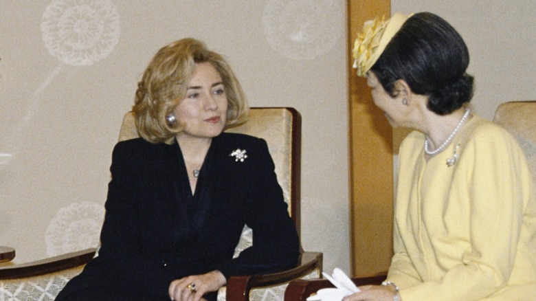 Dressed in a black suit, Hillary Clinton smiles lightly while engaging in a conversation