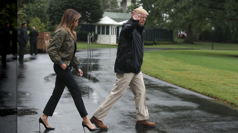 Dressed in a bomber jacket and an all-black outfit, Melania Trump walks behind a waving Donald Trump
