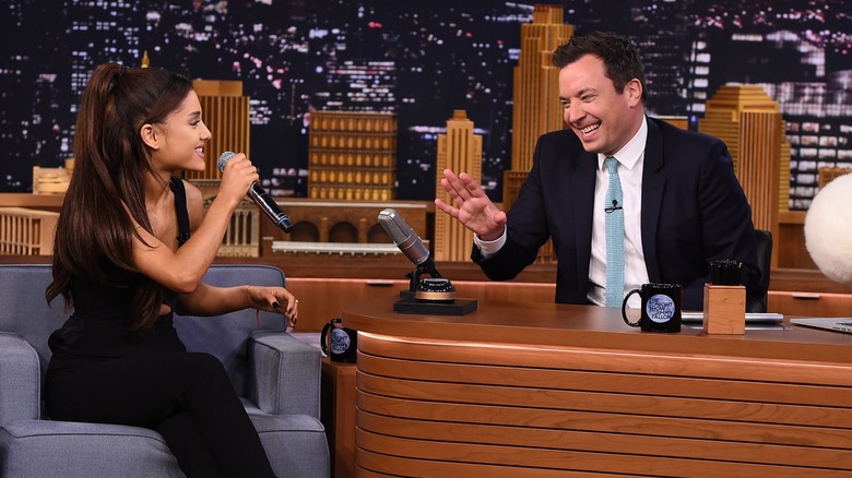 Ariana Grande speaks into a microphone as Jimmy Fallon makes a hand gesture while laughing.