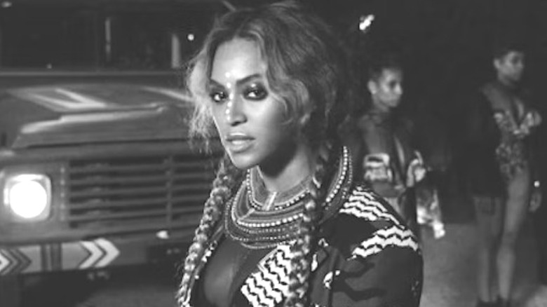 Beyoncé Knowles sporting African jewelry and a printed coat, with her dancers and a bus in the background.