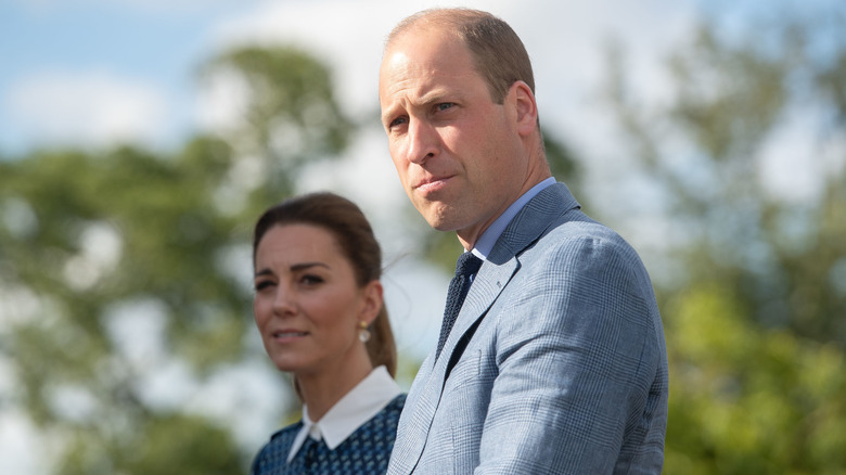 Kate Middleton standing behind a frowning Prince William