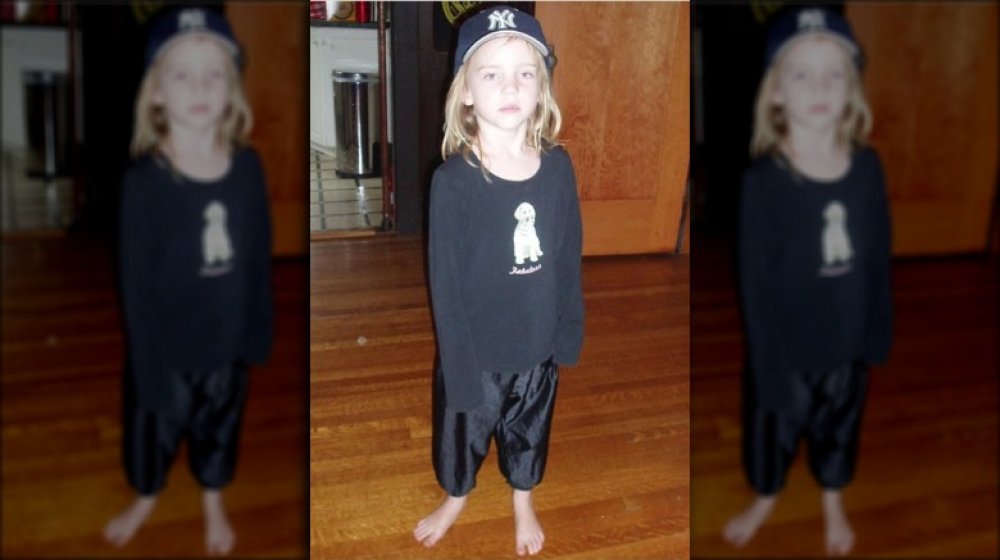 A young Billie Eilish wearing an all-black outfit, with a puppy shirt, shiny pants, and NY Yankees baseball cap