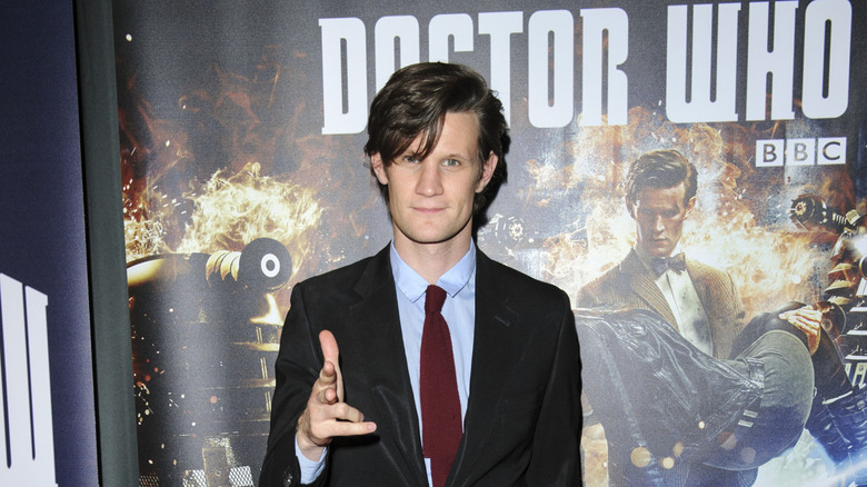Matt Smith wearing a black suit and maroon tie posing in front of a Doctor Who poster