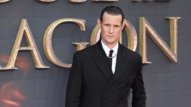Matt Smith with slicked back hair wearing a black suit coat, black tie, and gold chain on the red carpet