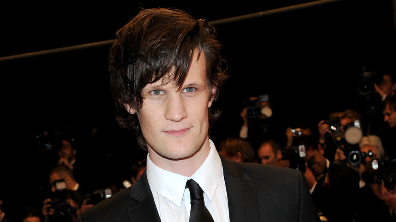 Matt Smith with long shaggy hair wearing a black suit and tie at a red carpet event