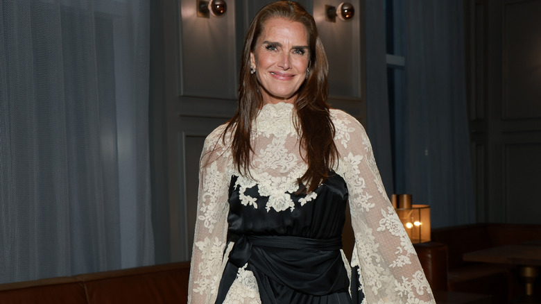 Brooke Shields smiling in a black and white dress with lace on the neckline, sleeves, and skirt.