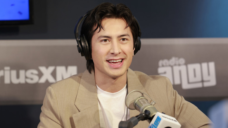 Hudson Williams wearing a tan suit jacket and white t-shirt with headphones on speaking into a radio mic