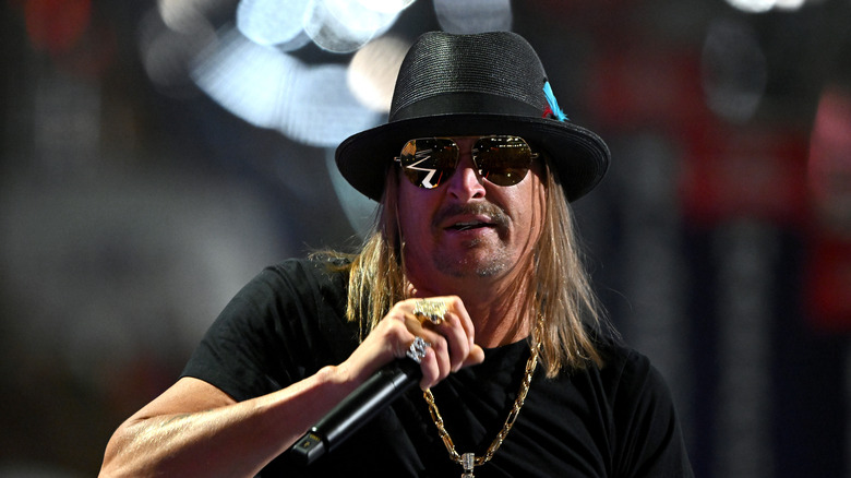 Kid Rock wearing sunglasses and a black hat while holding a microphone.