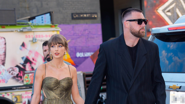 Taylor Swift in gold dress walking while holding hands with Travis Kelce in a black suit