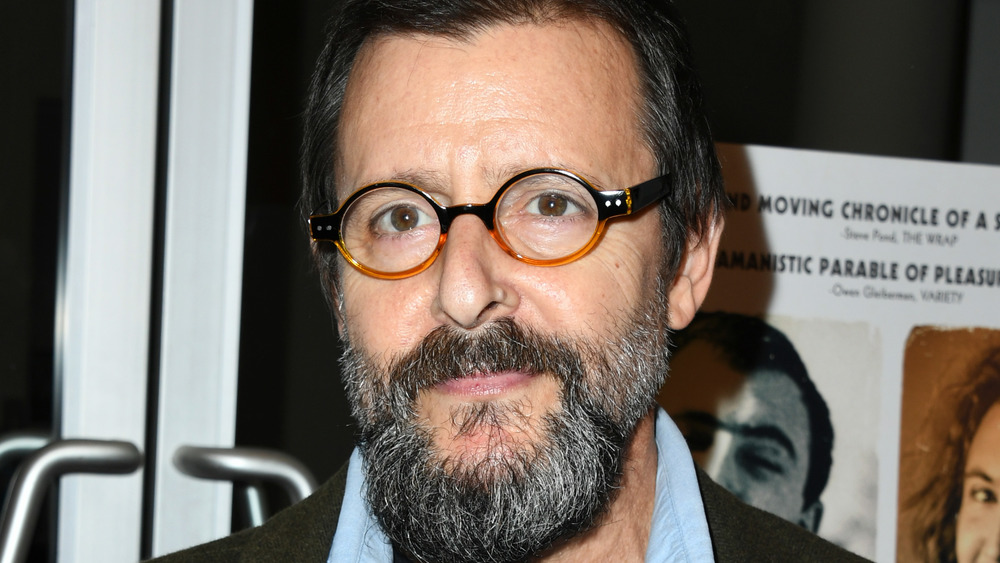 A bearded Judd Nelson smirking and wearing glasses 