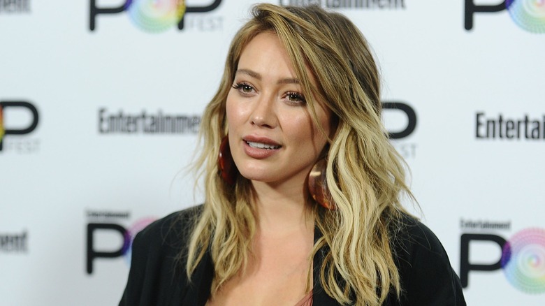 Hilary Duff stares blankly away from the camera while wearing a black coat and large brownish red earrings