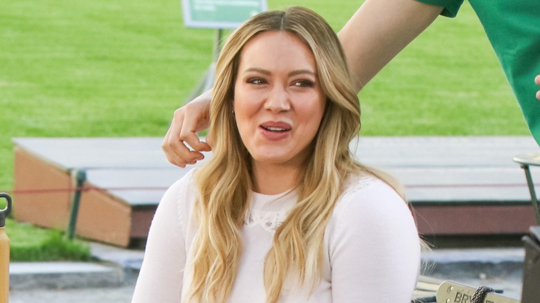 A seated Hilary Duff looks away from the camera while making a face