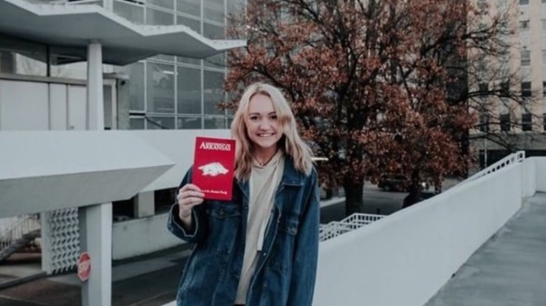 Wearing an oversized jacket, Paige Drummond smiles as she displays a red booklet.