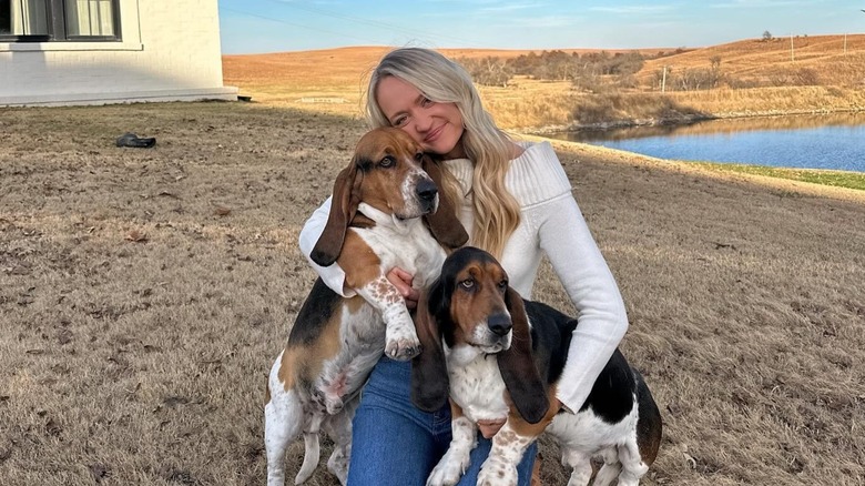 A smiling Paige Drummond cuddles her pet dogs as she kneels on dry grass.