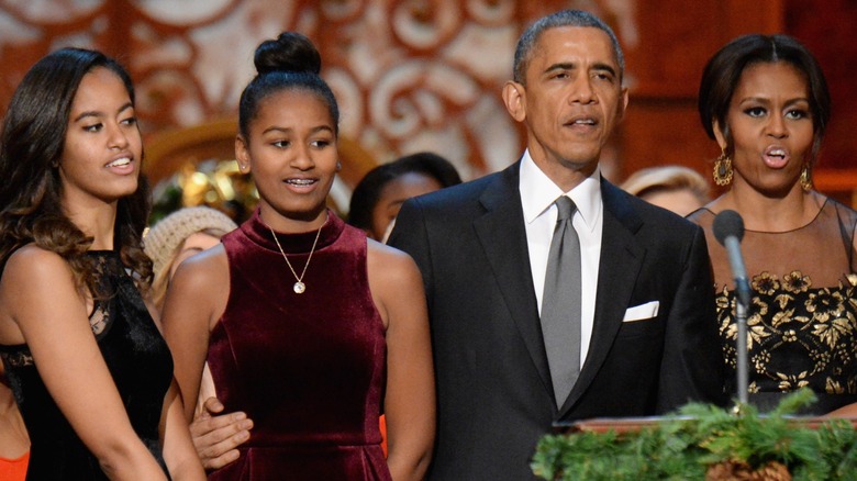 The Obama family stand in line and sing into a microphone, as Barack Obama wraps his arm around Sasha Obama.