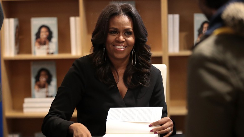 Michelle Obama smiles as she signs a copy of her book "Becoming," all while a reader watches on.