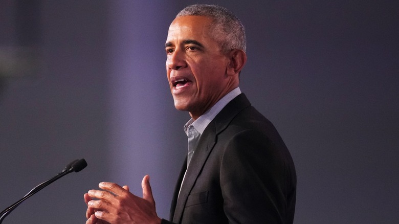 Dressed in a gray shirts and a black suit, Barack Obama speaks into a microphone.
