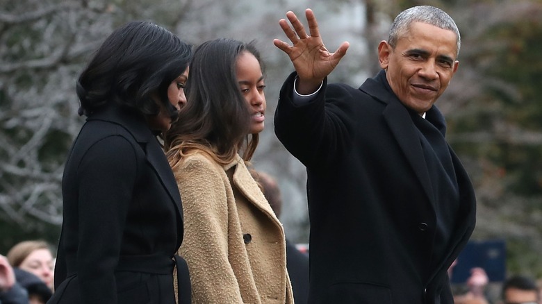 Barack Obama waves while Malia and Michelle Obama walk behind him.
