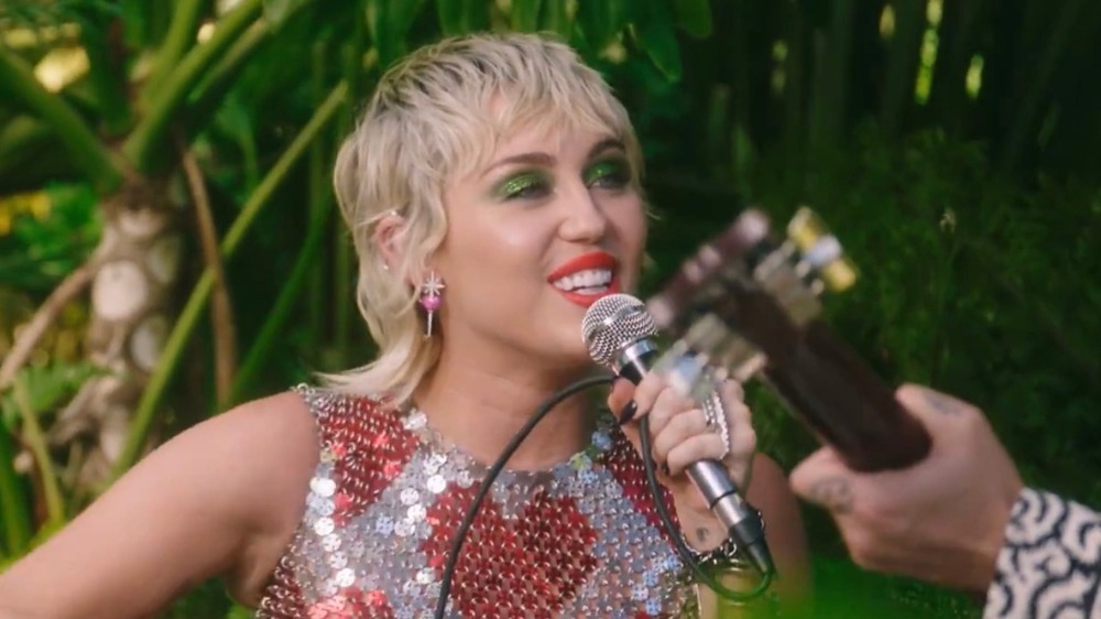 Miley Cyrus sings outdoors into a microphone with a guitarist's hand in view