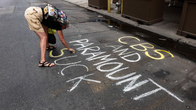 A woman with a backpack writes "CBS and Paramount Suck" in chalk on the street