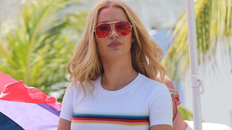 Wearing a set of sunglasses and a white shirt with a rainbow strip, Iggy Azalea makes a serious face for the camera.