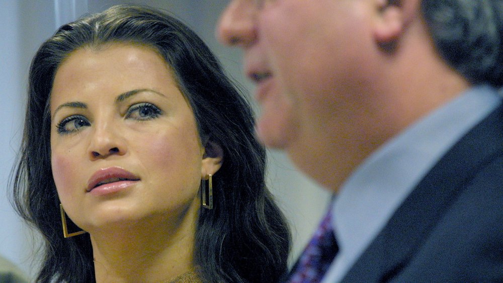 Yasmine Bleeth wearing a beige turtleneck, full makeup, and gold rectangle earrings, glancing to her left with a serious expression