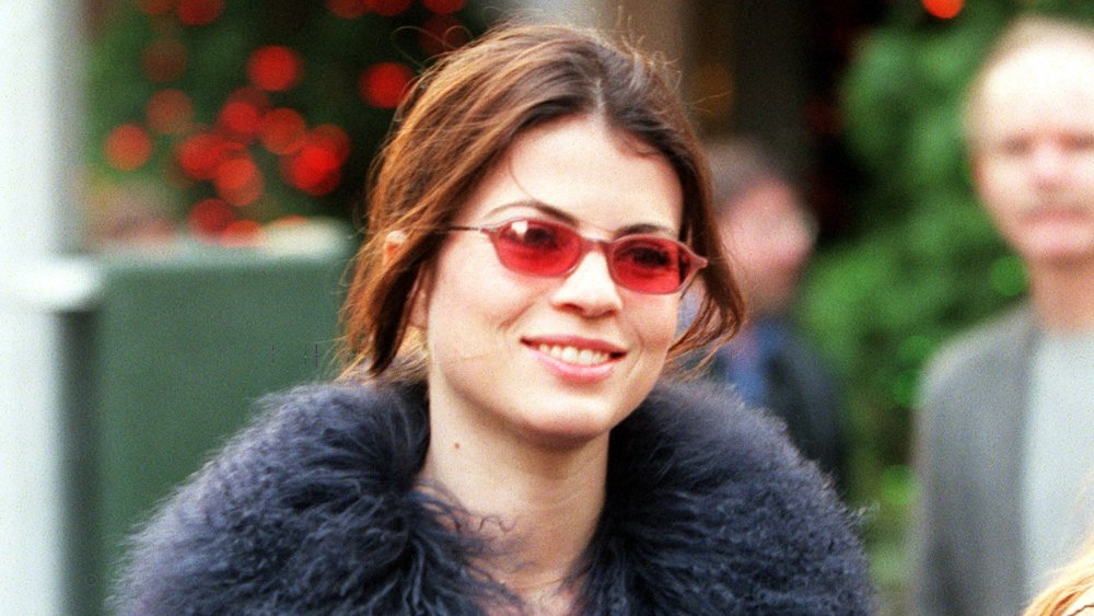 Yasmine Bleeth smiling and walking, wearing red-tinted glasses and a black, fuzzy coat